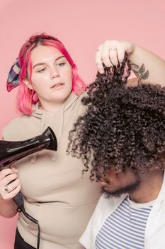 Concentrated plump female hairdresser drying black Afro hair of African American male client on pink background in modern light studio