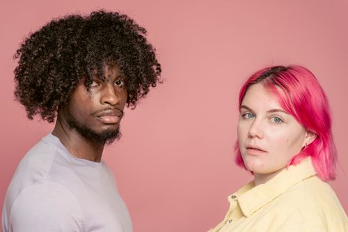 Free Serious African American man near woman with dyed hair looking at camera while standing on pink background in light studio Stock Photo