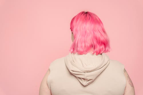 Back view of faceless female with dyed hair wearing sleeveless shirt standing against pink background
