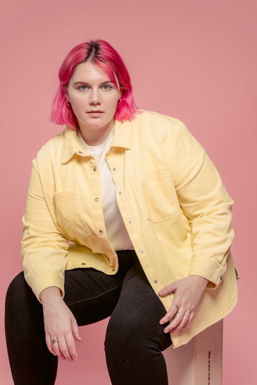 Confident young female plus size model wearing casual clothes sitting against pink background and looking at camera