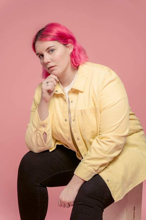 Young female model with dyed bright hair wearing casual clothes sitting against pink background and touching chin