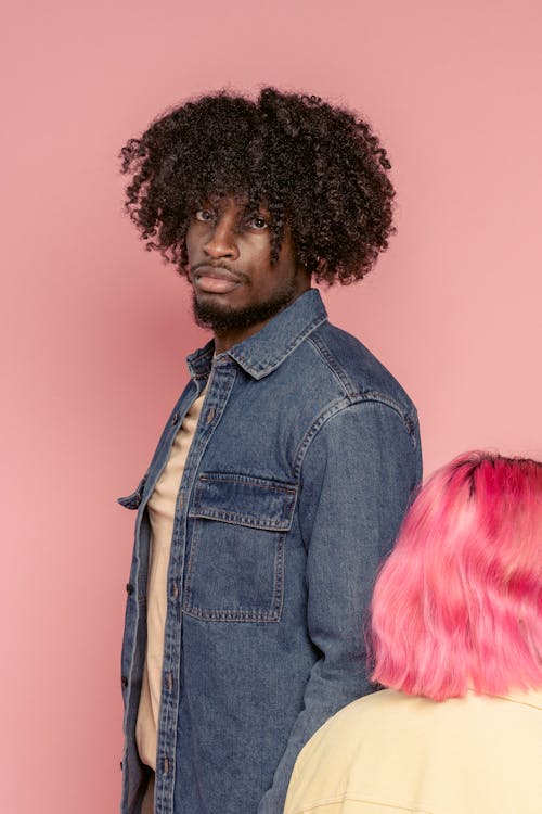 Confident African American male with curly hair wearing denim jacket standing near crop woman against pink background