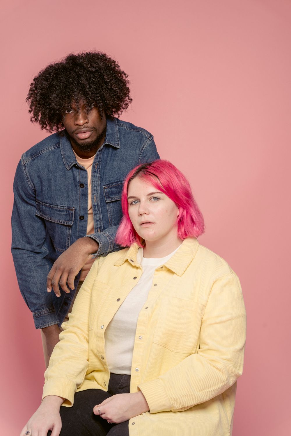 Two models in a denim jacket and butter-yellow overshirt against a pink studio backdrop — photo by John Diez on Pexels