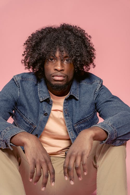 Unemotional African American male with Afro hairstyle wearing casual clothes sitting on floor against pink background