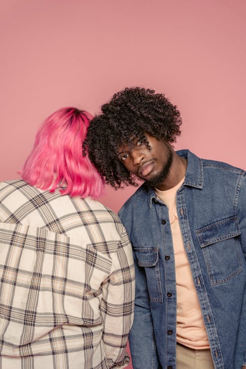 Serious African American man with curly hair wearing casual clothes bending to girlfriend with bright dyed hair while standing against pink background
