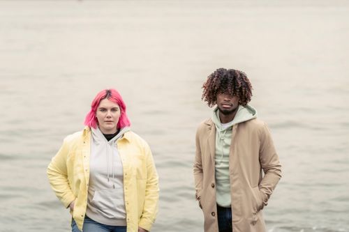 Free Serious woman and African American man in outerwear looking at camera while standing with hands in pockets on waterfront near rippling water Stock Photo