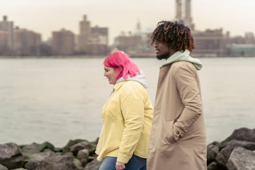 Two people walk along a riverbank, enjoying a casual and quiet day outdoors.