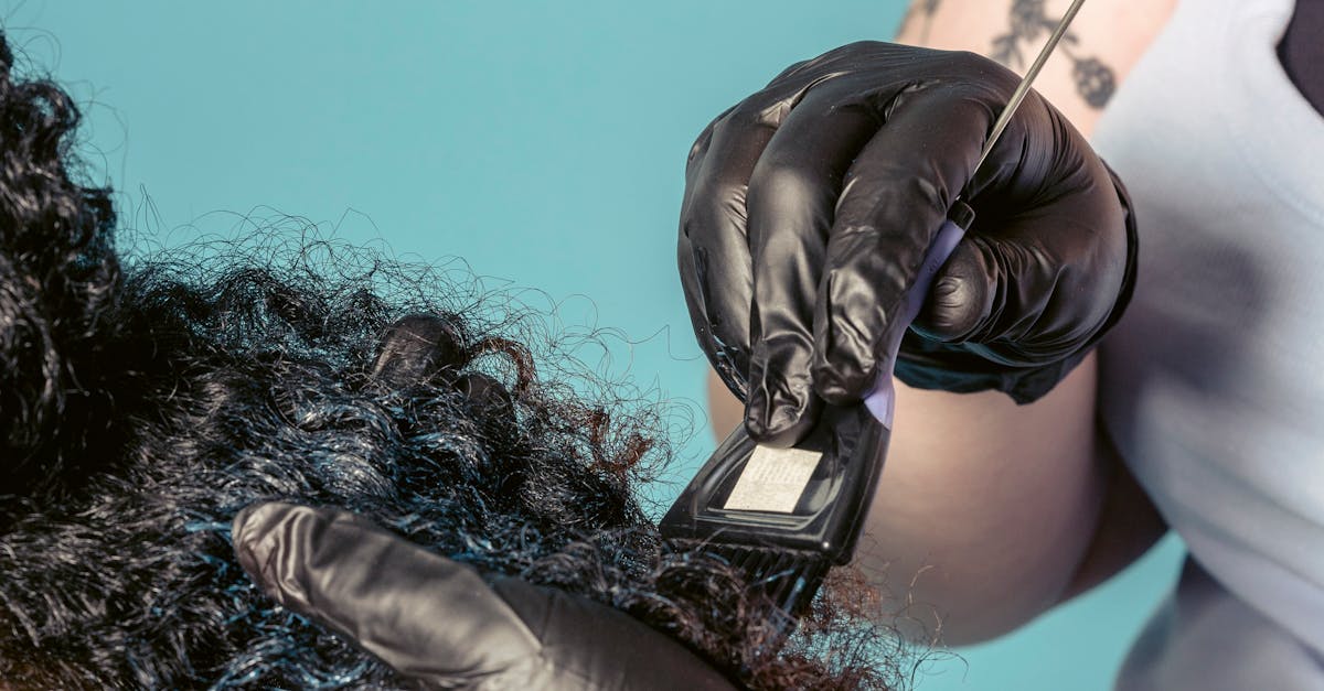 Crop anonymous female master with tattooed arms in latex gloves using brush to dye curly hair of client in salon