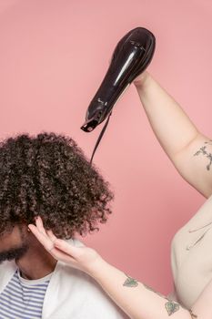 Faceless female hairstylist with tattoo drying curly hair of unrecognizable African American man with beard using hairdryer in studio salon against pink wall