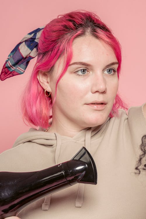 Headshot of focused young female hair dresser with pink wavy hair in sweatshirt holding air hair dryer while working with Afro hair of customer in salon