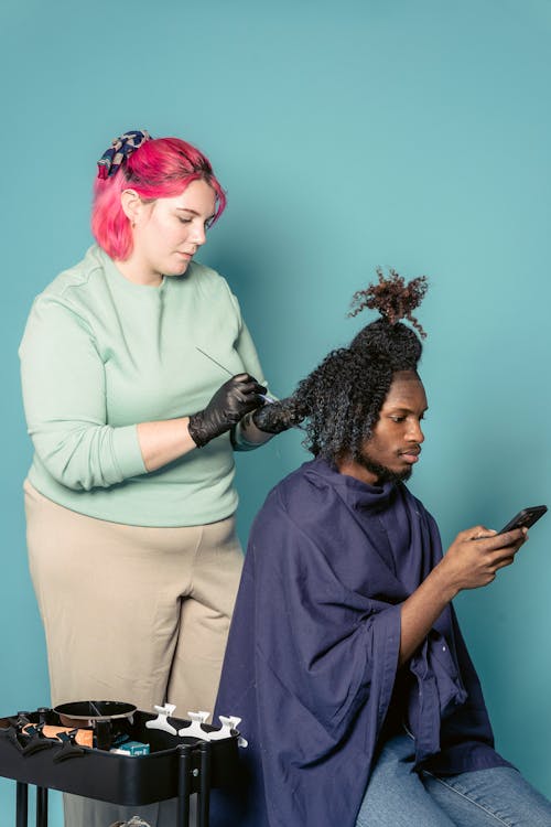 Side view of curvy hairstylist with pink hair wearing casual clothes applying coloring cream on hair with afro curls of African American customer with beard