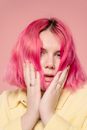 Tender young female with bright dyed hair touching cheeks while looking at camera with mouth opened