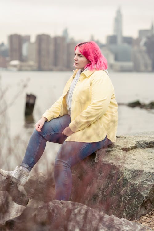 Full body lonely plump female with pink hair wearing casual clothes sitting on boulder on river shore and looking away thoughtfully