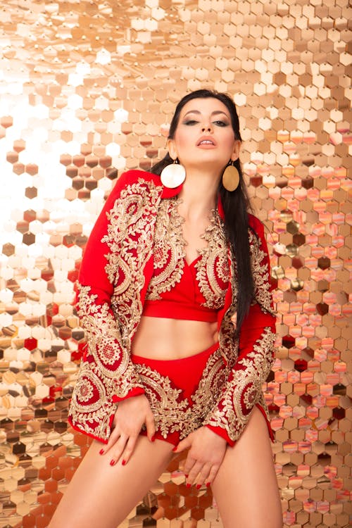 Charming female in stylish red clothes with bright makeup and accessories posing near glowing wall and looking at camera