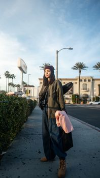 Full body of calm young woman in trendy outfit standing with ukulele in case on shoulder on asphalt roadway in city in sunny day