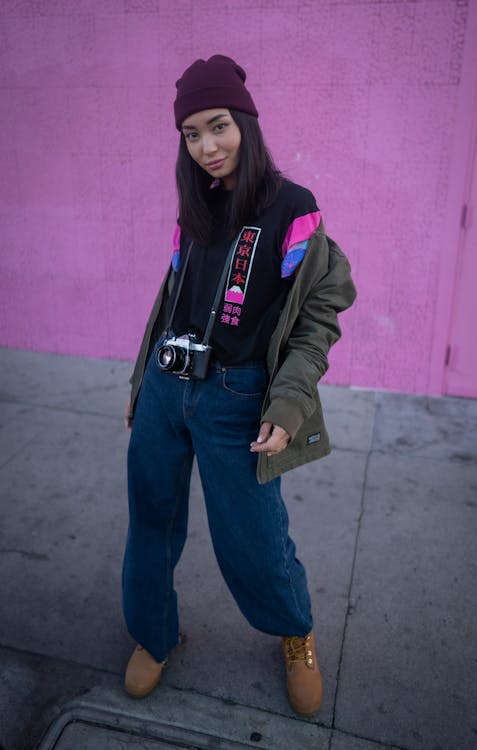 Full body of young woman in stylish clothes standing with photo camera on asphalt road and looking at camera against pink wall of building