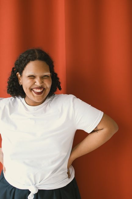 Happy woman with curly hair posing in a white shirt against a red background, exuding confidence and joy.