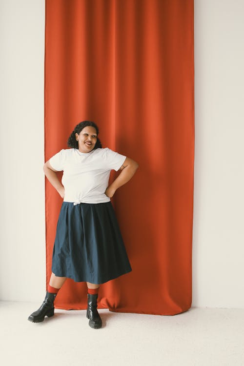 A Woman in White Shirt and Black Skirt Standing Near the Red Curtain with Her Hands on Her Waist
