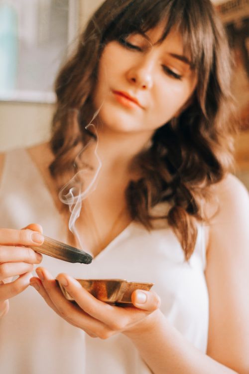 Free Crop young female with long wavy hair in white blouse burning aromatic sandal stick on blurred background Stock Photo