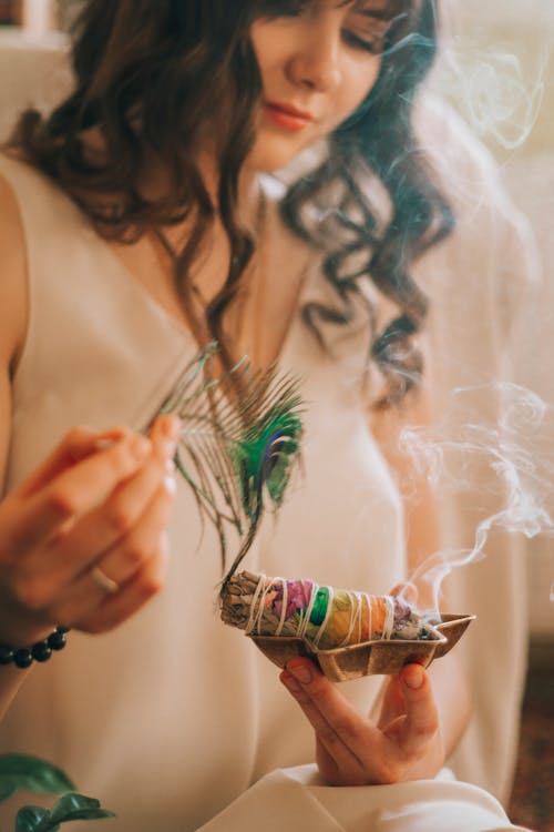 Free Crop female with wavy dark hair smiling and smelling smoke of smudge sticks on blurred background Stock Photo