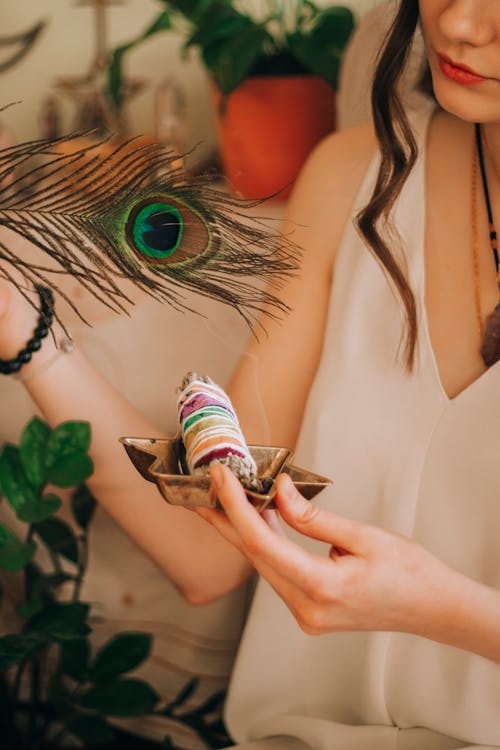 Free Crop anonymous female in white blouse with smudge timber sticks and colorful peacock feather Stock Photo