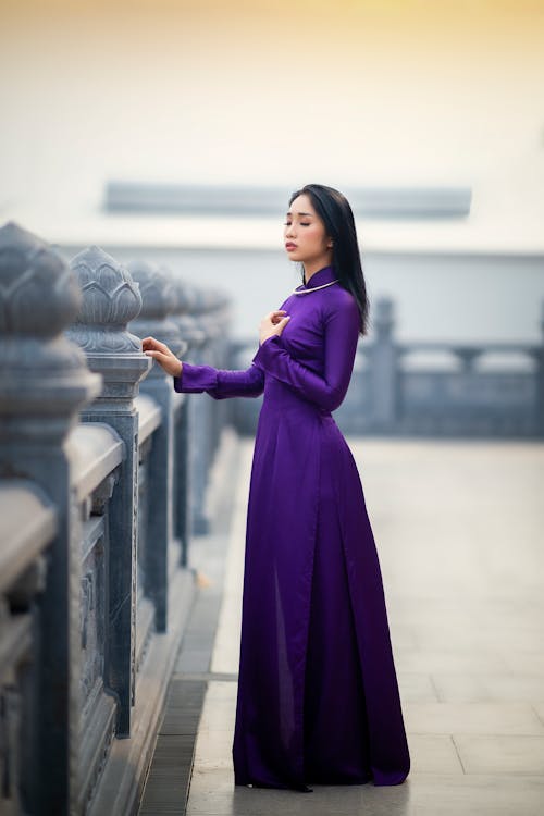 Thoughtful young Asian woman in violet long dress standing on bridge with closed eyes