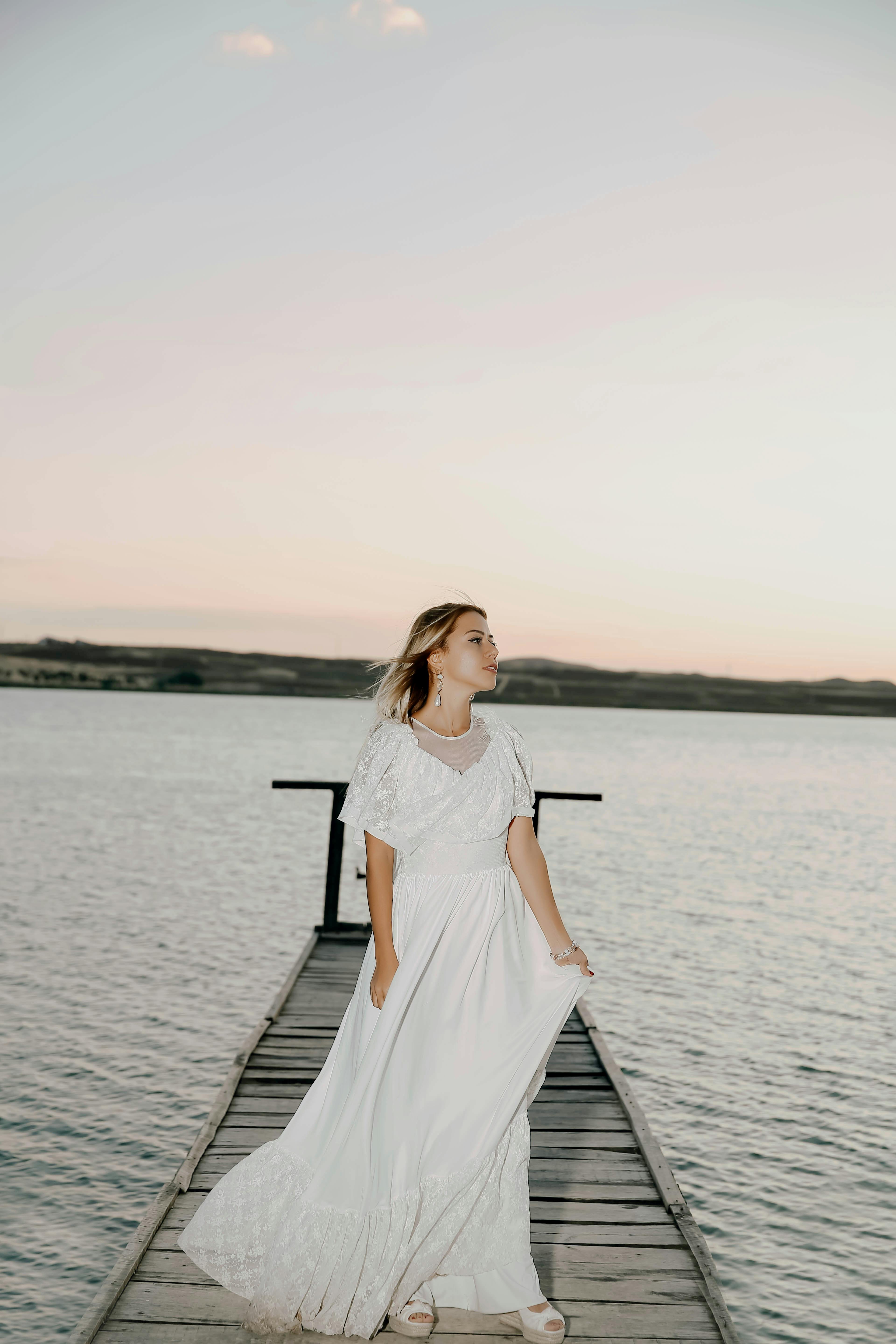 A serene image of a woman in a flowing white dress standing on a lakeside dock during sunset.