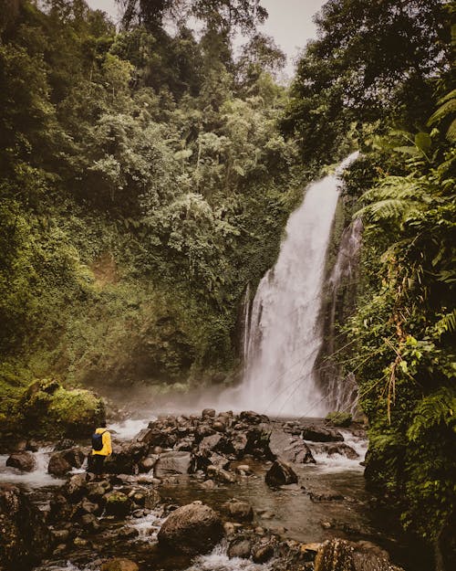 Person in Yellow Hoodie and Backpack Standing on Rock Near Waterfalls