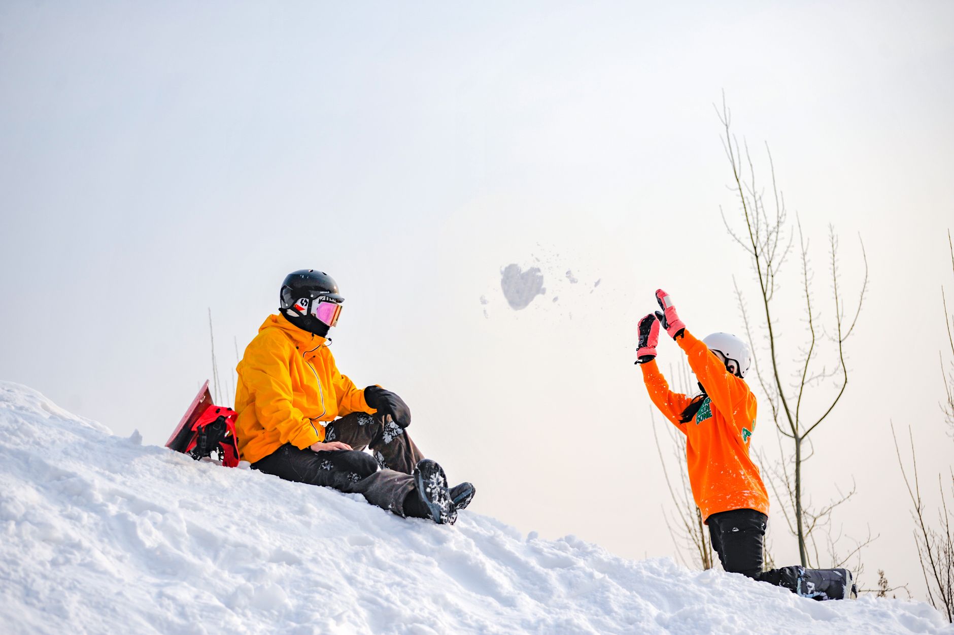 Two people dressed in winter gear enjoying a snow-covered hillside, throwing snow.