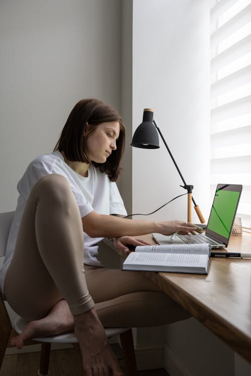 Young female student in casual clothes reading book and typing on laptop while preparing for exams sitting at table at home