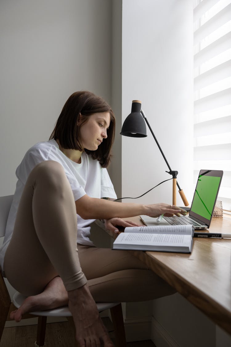 Young Woman Reading Book And Using Laptop At Home