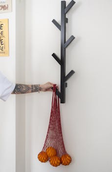 A tattooed hand holds a red net bag filled with oranges, hanging on a modern wall hook.