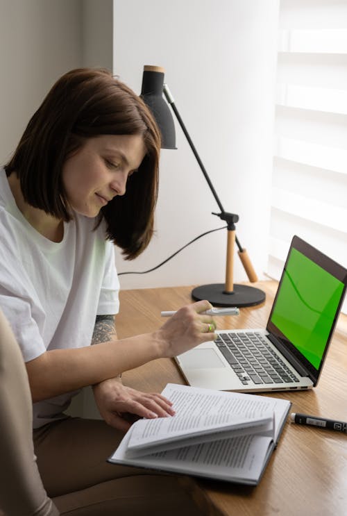 Free Concentrated young female freelancer in casual clothes reading book and working remotely on laptop with chromakey screen sitting at table at home Stock Photo