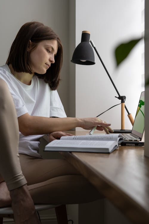 Free Young female freelancer working online on laptop and reading book Stock Photo