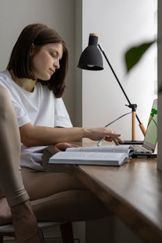 Young woman focusing on study materials at home, utilizing a laptop and book under a desk lamp.