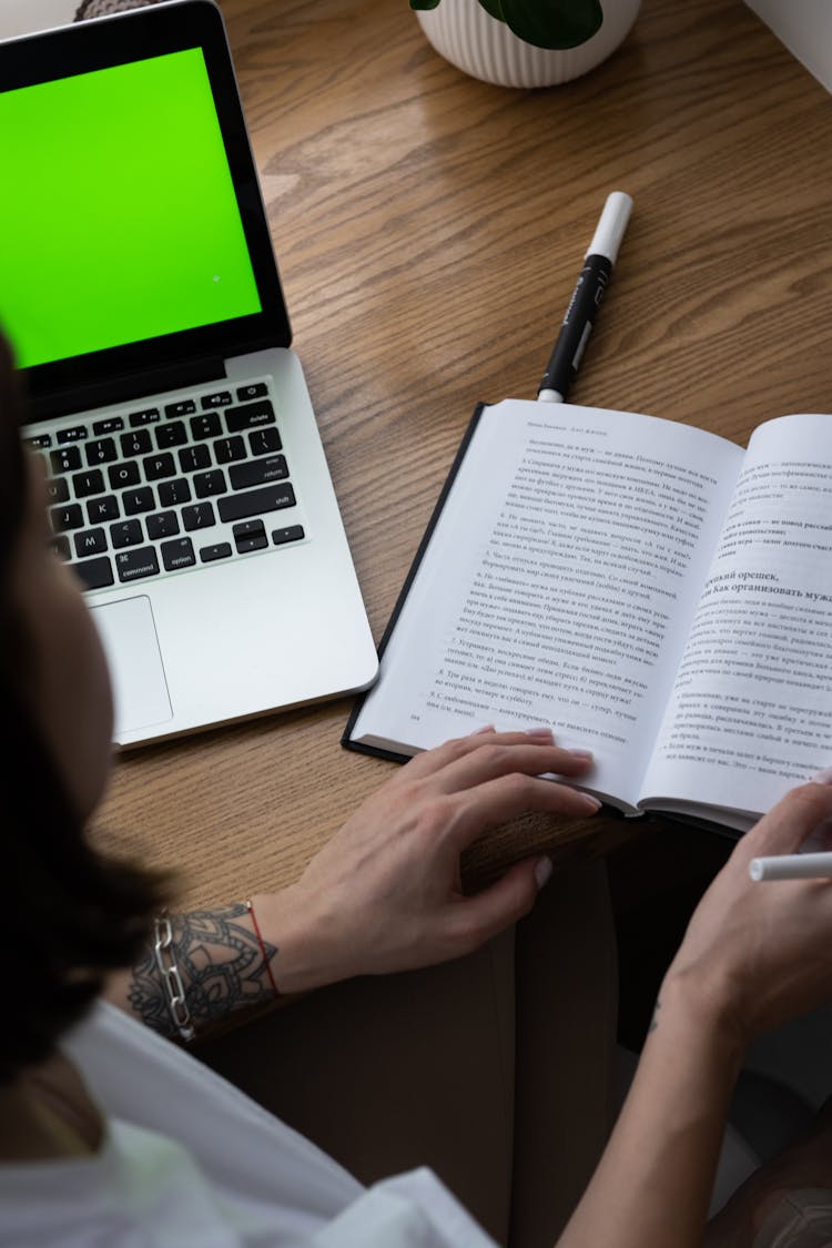 Anonymous Woman Reading Book While Studying Remotely At Table With Laptop