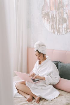 Asian woman in bathrobe using laptop on bed, highlighting cozy indoor lifestyle.