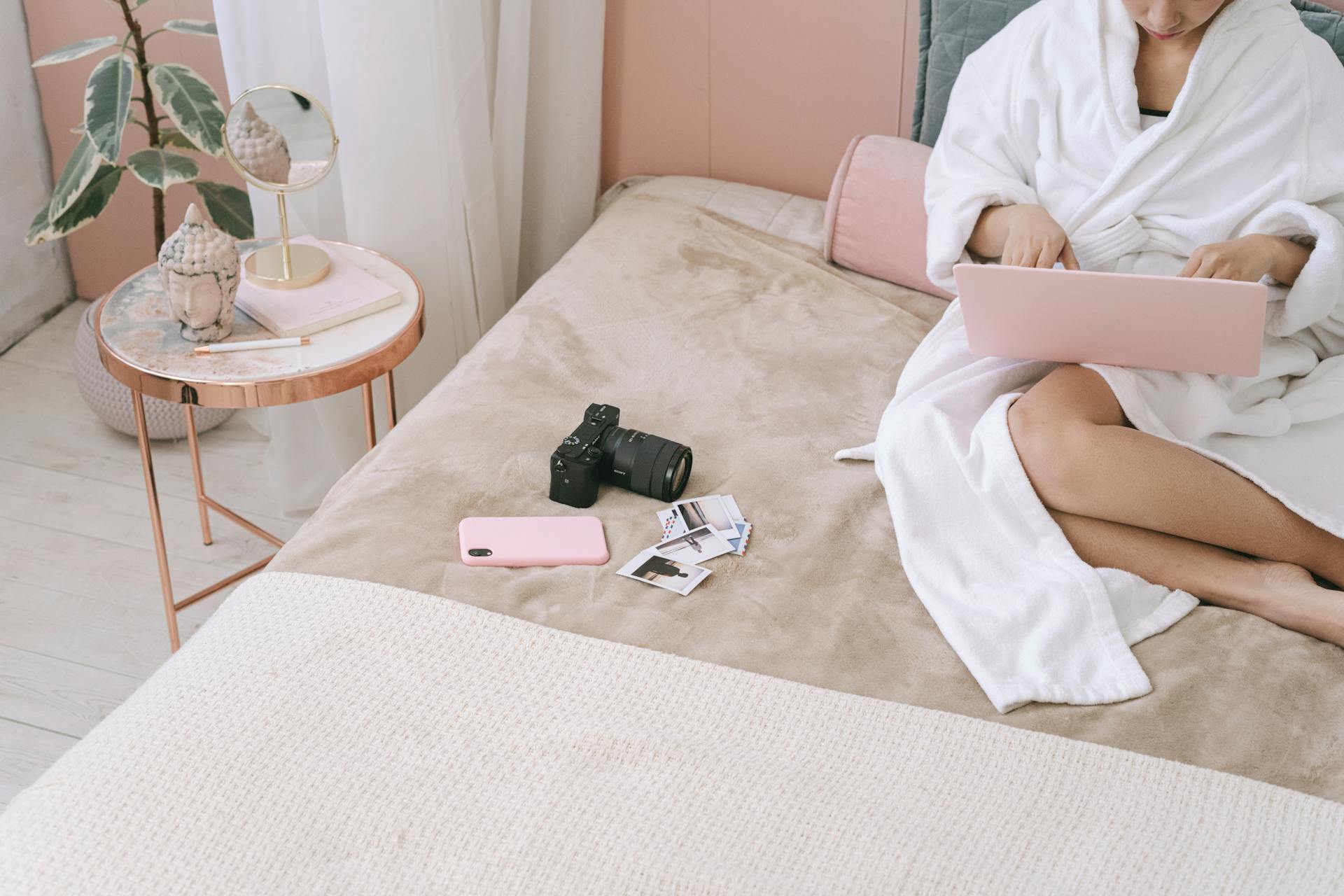 A Person in White Robe Sitting on Bed with Camera and Cellphone