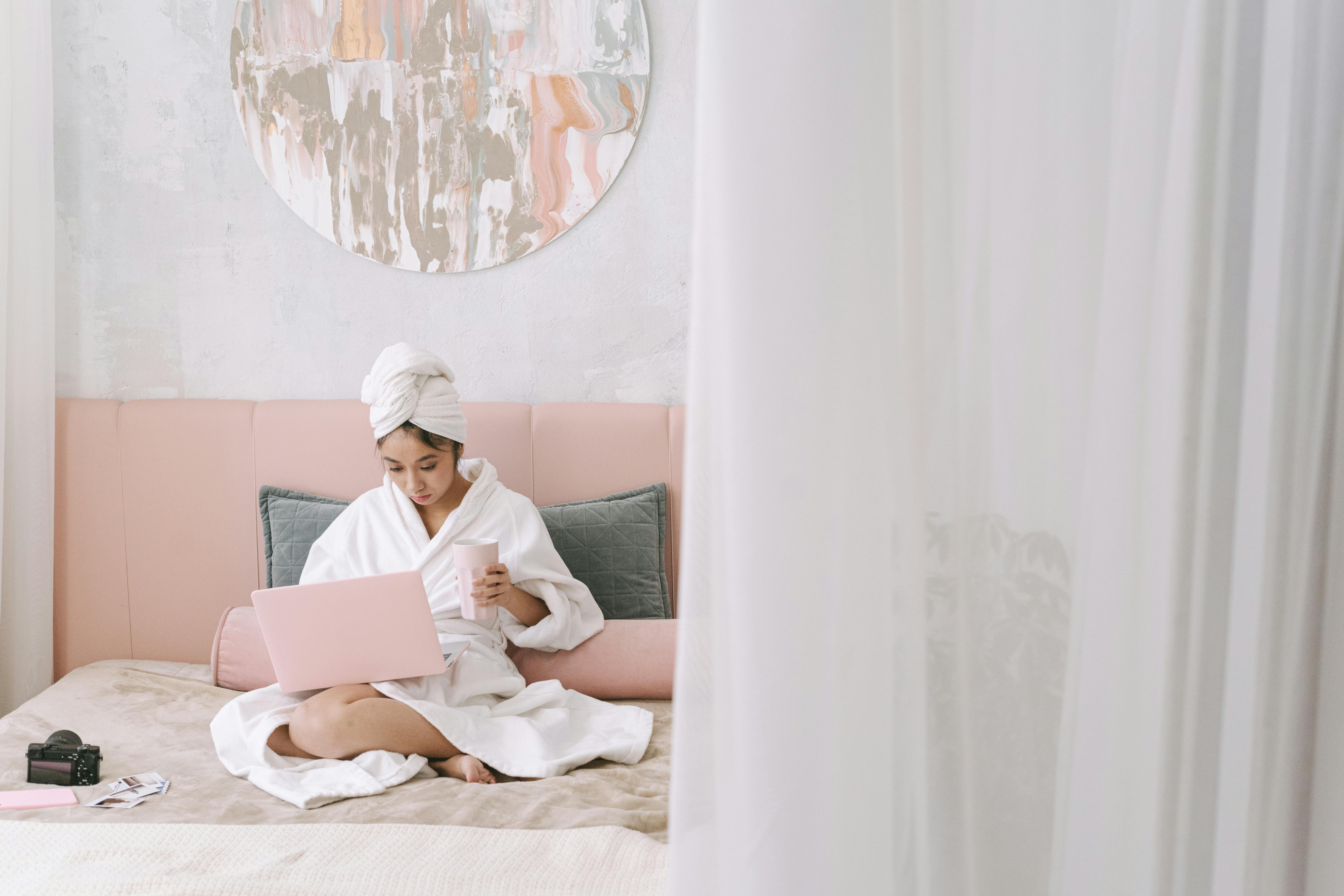 An Asian woman in a bathrobe using a laptop on her bed, exuding a cozy morning vibe.
