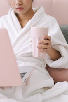 Woman relaxing in a white robe, holding a pink mug and using a laptop indoors.