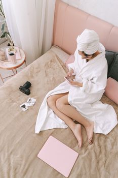 A woman in a robe sits on a bed using a smartphone, with a camera and laptop nearby.