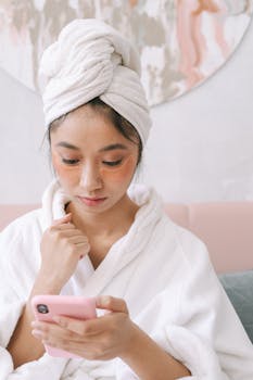 A woman in a bathrobe and towel uses her phone while wearing under-eye patches for beauty care.