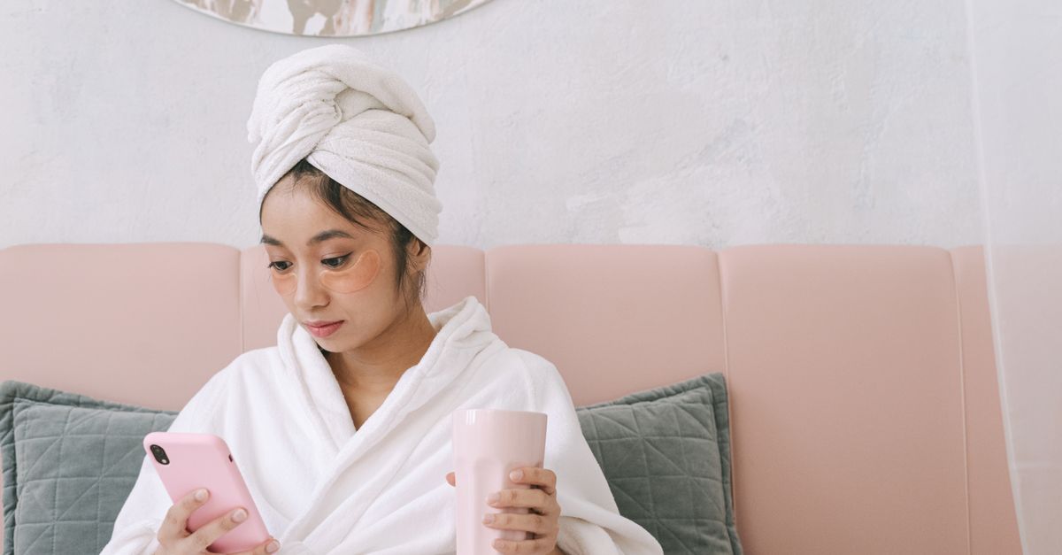 Woman in bathrobe with under-eye masks holding phone and mug, embodying relaxation and beauty.