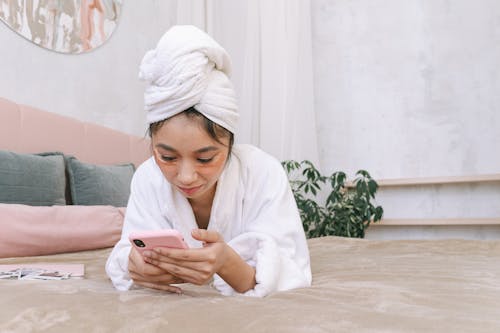 A Woman in a White Bathrobe With a White Towel Wrapped Around Her Head Holding and Looking at a Smartphone