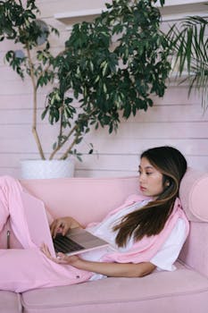 Young woman in pink clothing working on a laptop from home, lying on a pink sofa in a cozy setting.