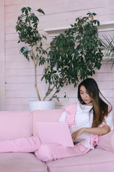 Young woman in pink outfit working on a laptop, relaxing on a cozy sofa with green plants in the background.