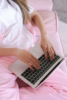 A young woman in pink pants works on her laptop from a cozy home setting. Perfect representation of remote work lifestyle.