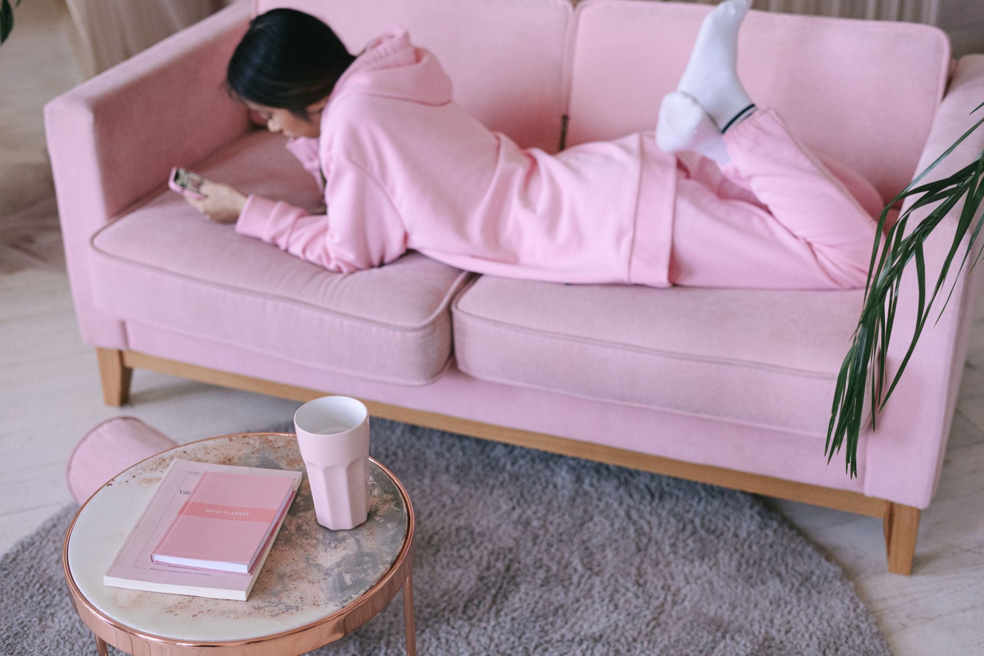 A woman in pink loungewear lying on a pink sofa, using a smartphone in a cozy apartment.
