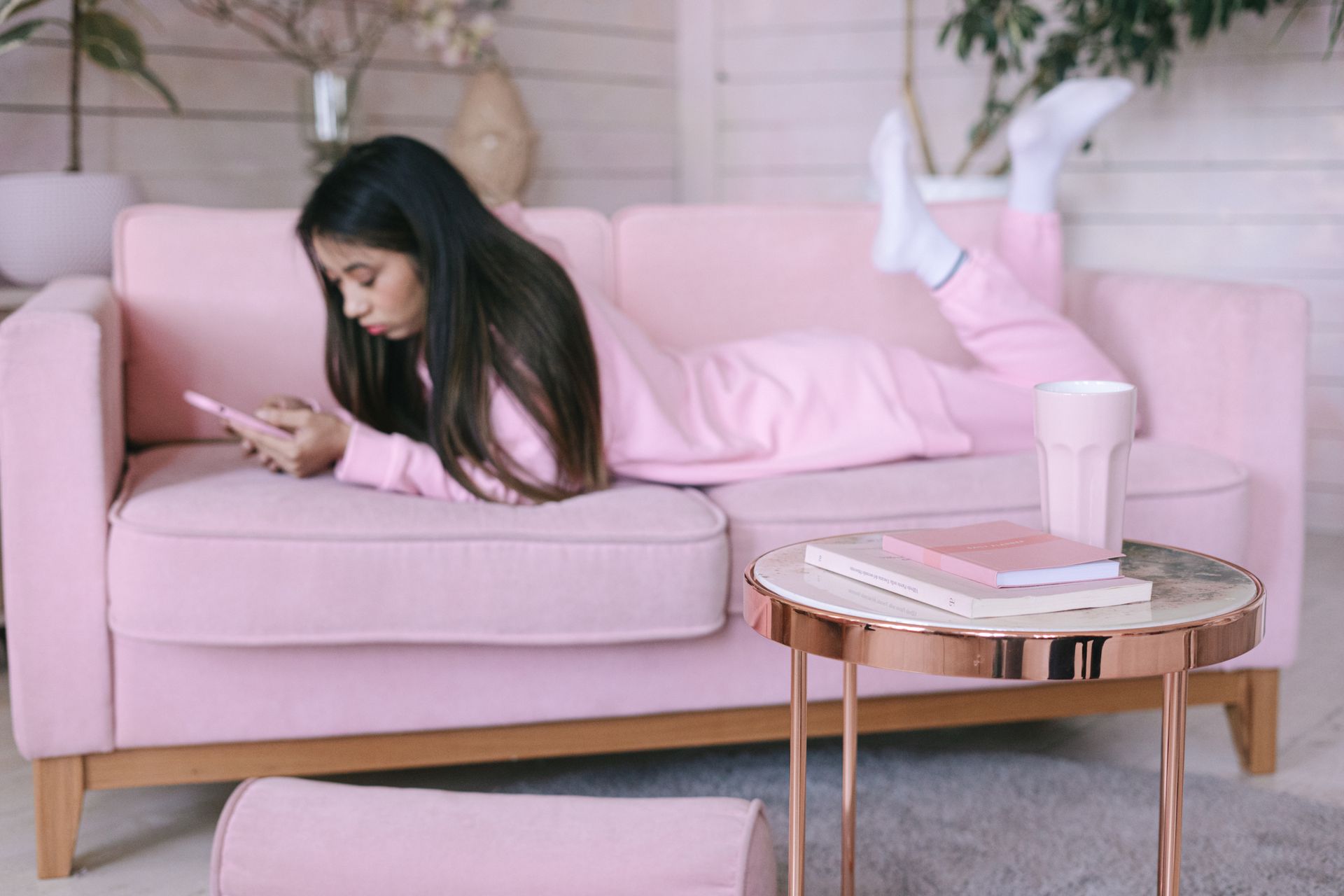 A young adult woman in pink loungewear relaxes on a sofa using her smartphone, embodying a cozy indoor lifestyle.