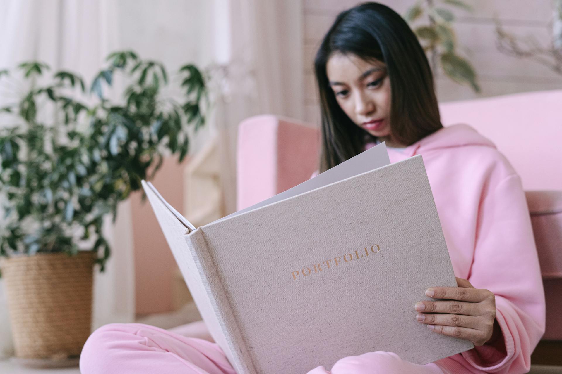 Asian teenager in pink hoodie reading a portfolio indoors with potted plants.